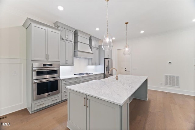 kitchen featuring hanging light fixtures, stainless steel appliances, light hardwood / wood-style flooring, an island with sink, and custom exhaust hood