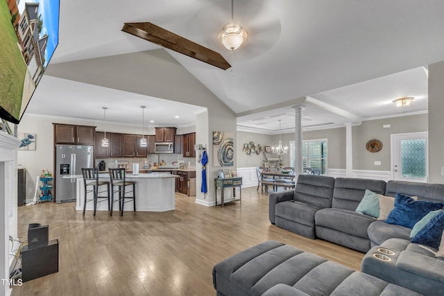 living room featuring ceiling fan with notable chandelier, ornate columns, light hardwood / wood-style floors, ornamental molding, and lofted ceiling with beams