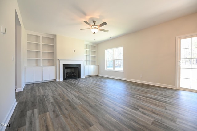 unfurnished living room with dark wood-type flooring, a wealth of natural light, ceiling fan, and built in shelves