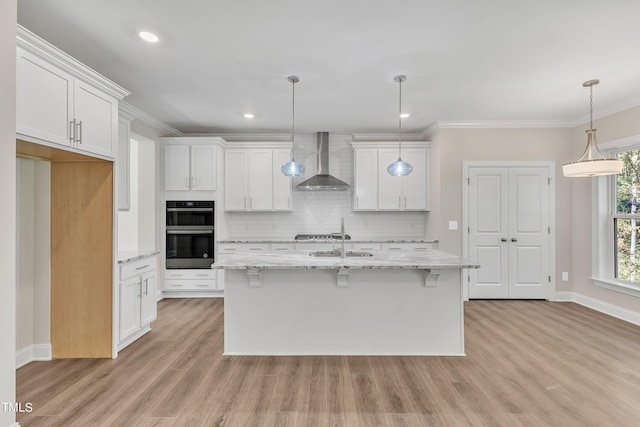 kitchen with wall chimney range hood, double oven, white cabinetry, crown molding, and light hardwood / wood-style flooring