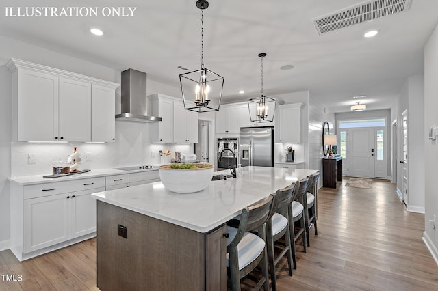 kitchen with wall chimney range hood, light hardwood / wood-style flooring, stainless steel fridge, a kitchen island with sink, and white cabinets