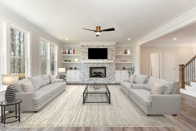 living room featuring ceiling fan, crown molding, built in features, light hardwood / wood-style flooring, and a stone fireplace