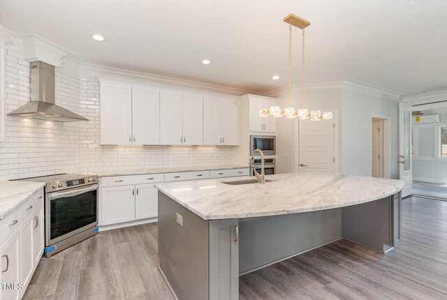 kitchen featuring hardwood / wood-style floors, a kitchen island with sink, wall chimney range hood, appliances with stainless steel finishes, and decorative light fixtures