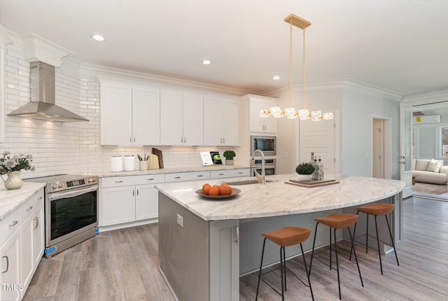 kitchen featuring wall chimney exhaust hood, an island with sink, pendant lighting, white cabinets, and appliances with stainless steel finishes