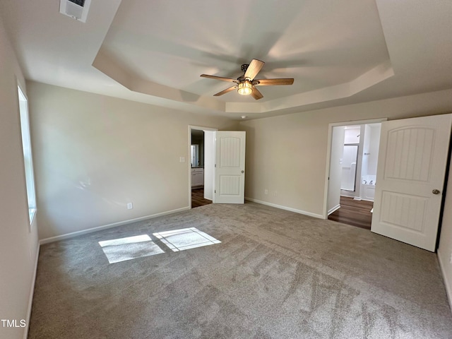 unfurnished bedroom featuring ceiling fan, ensuite bathroom, a raised ceiling, and dark colored carpet