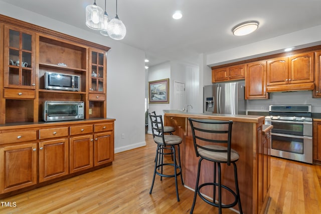 kitchen featuring light wood-style flooring, glass insert cabinets, brown cabinets, stainless steel appliances, and under cabinet range hood