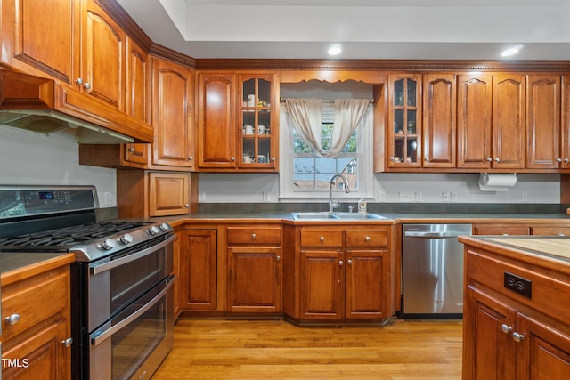 kitchen with brown cabinets, light wood finished floors, appliances with stainless steel finishes, glass insert cabinets, and a sink