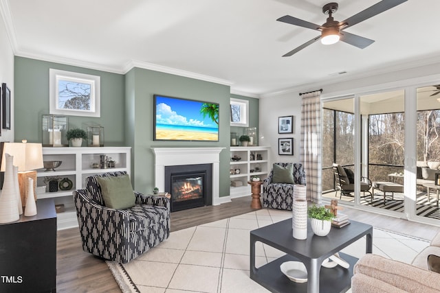 tiled living room with ceiling fan, visible vents, crown molding, and a glass covered fireplace