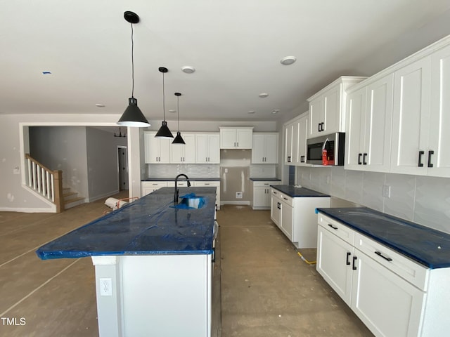 kitchen featuring a kitchen island with sink, a sink, white cabinetry, backsplash, and stainless steel microwave