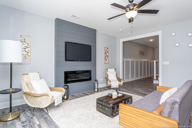 living area featuring a large fireplace, a ceiling fan, baseboards, and wood finished floors