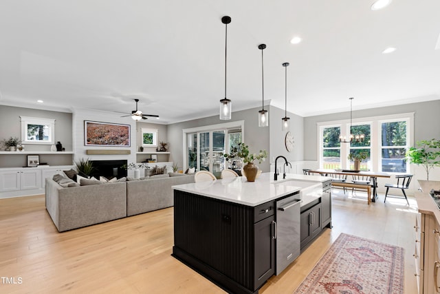 kitchen with a kitchen island with sink, sink, ceiling fan with notable chandelier, a large fireplace, and stainless steel dishwasher