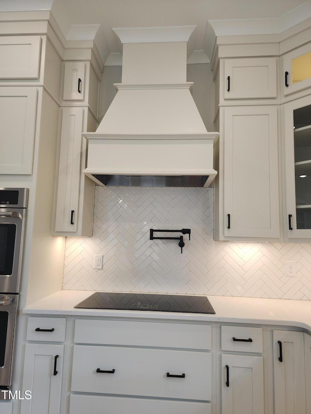 kitchen with double oven, black electric cooktop, decorative backsplash, glass insert cabinets, and custom range hood