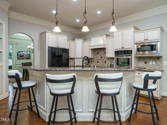 kitchen with pendant lighting, custom exhaust hood, dark wood-type flooring, appliances with stainless steel finishes, and a center island with sink