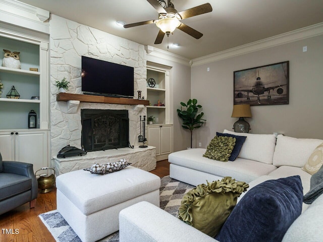 living room with crown molding, wood-type flooring, a stone fireplace, built in features, and ceiling fan