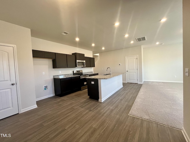 kitchen with hardwood / wood-style floors, sink, light stone countertops, an island with sink, and appliances with stainless steel finishes