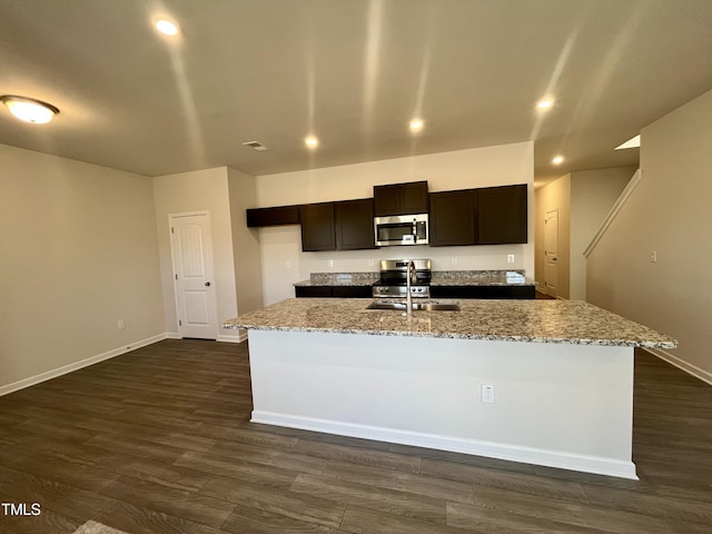 kitchen with sink, dark wood-type flooring, stainless steel appliances, light stone counters, and an island with sink