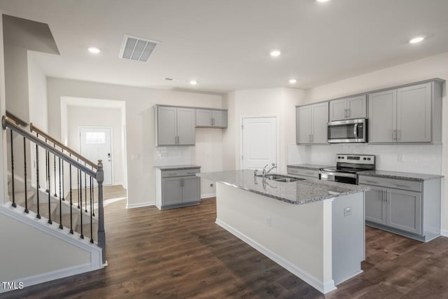 kitchen featuring a center island with sink, appliances with stainless steel finishes, light stone countertops, dark hardwood / wood-style floors, and gray cabinetry