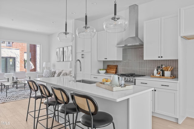 kitchen with white cabinetry, wall chimney exhaust hood, pendant lighting, a center island with sink, and light wood-type flooring