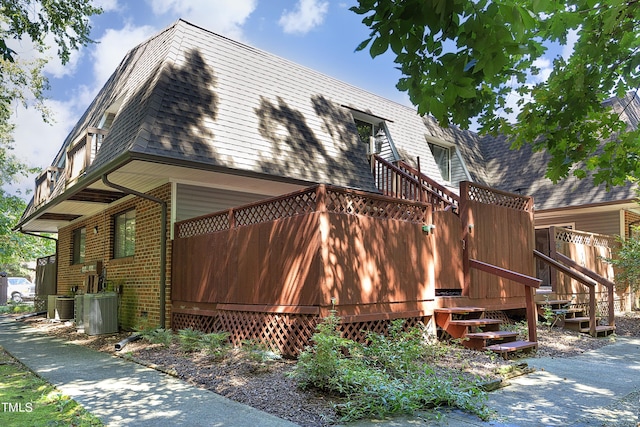 view of side of home with mansard roof, cooling unit, brick siding, and roof with shingles