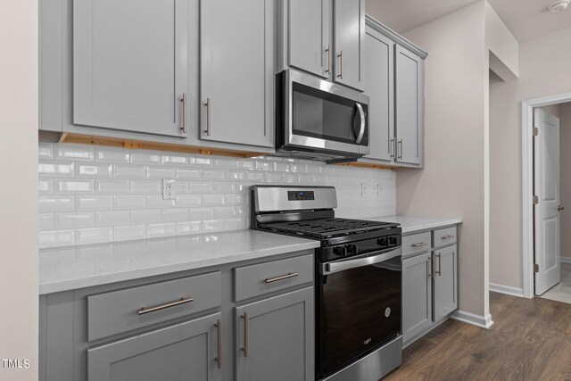 kitchen with gray cabinetry, backsplash, light stone counters, stainless steel appliances, and dark hardwood / wood-style floors