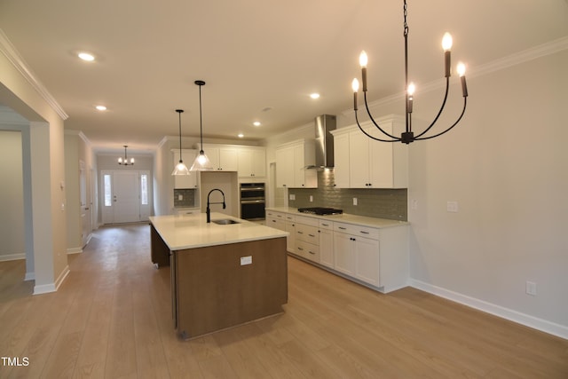 kitchen with an inviting chandelier, a center island with sink, wall chimney range hood, decorative light fixtures, and white cabinetry