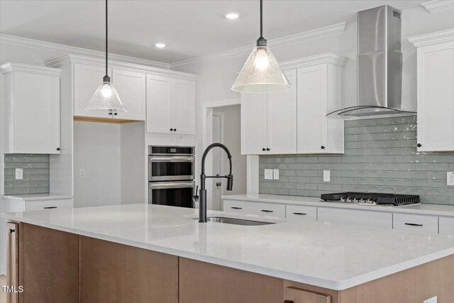 kitchen with a kitchen island with sink, stainless steel appliances, wall chimney range hood, white cabinetry, and pendant lighting