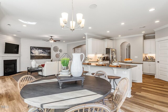 dining space featuring arched walkways, ceiling fan with notable chandelier, light wood-style floors, a glass covered fireplace, and crown molding