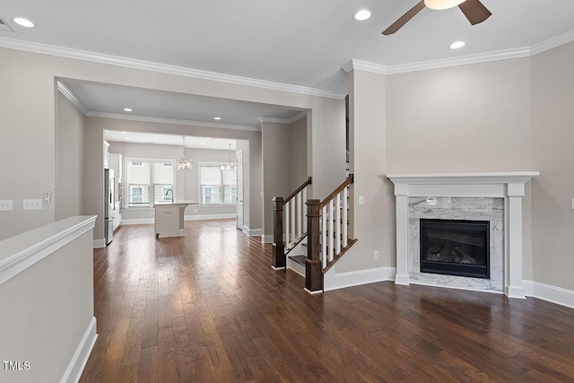 unfurnished living room featuring stairway, a premium fireplace, ceiling fan with notable chandelier, baseboards, and dark wood-type flooring
