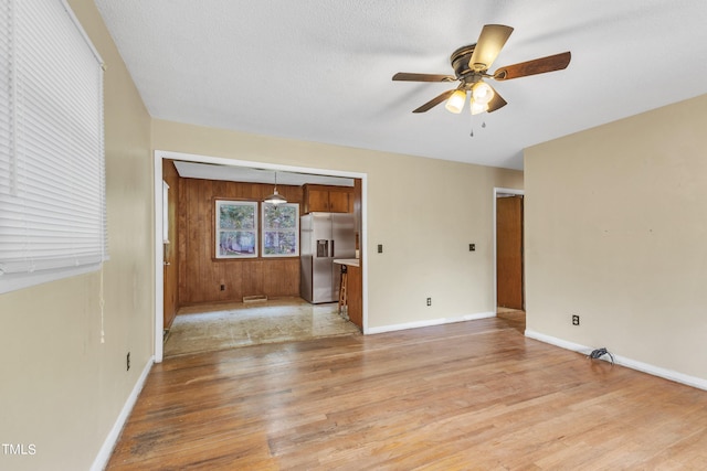 unfurnished living room featuring a textured ceiling, wooden walls, light wood finished floors, baseboards, and ceiling fan