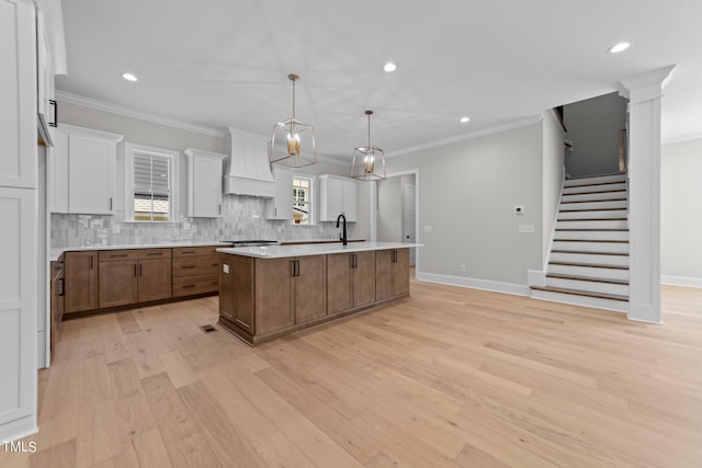 kitchen featuring light wood-type flooring, white cabinetry, custom range hood, a center island with sink, and decorative light fixtures