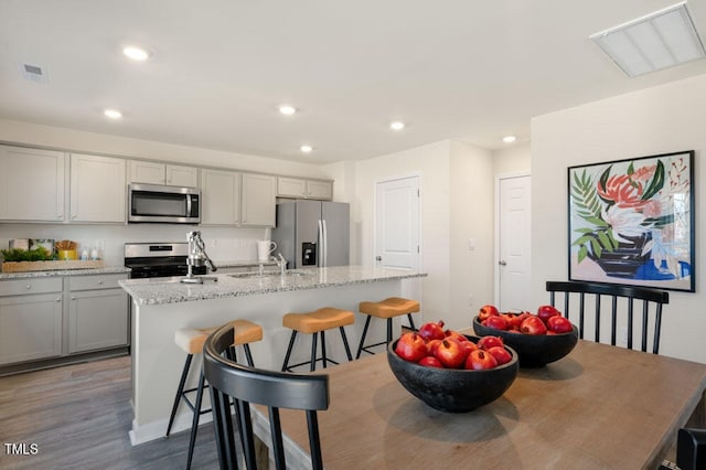 kitchen with hardwood / wood-style floors, stainless steel appliances, an island with sink, light stone countertops, and sink