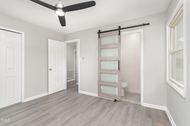 unfurnished bedroom with ensuite bathroom, a barn door, light wood-type flooring, ceiling fan, and multiple windows