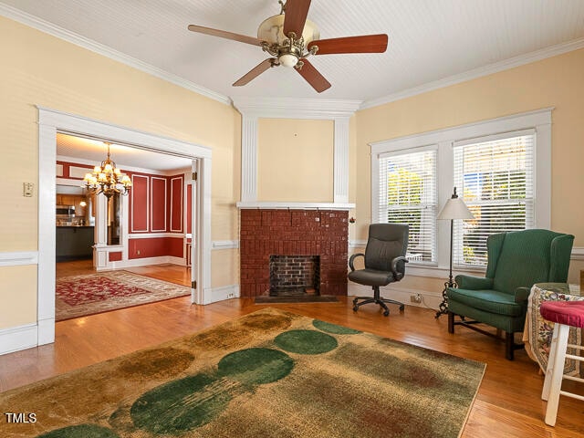 living area featuring ornamental molding, ceiling fan with notable chandelier, a brick fireplace, and wood-type flooring