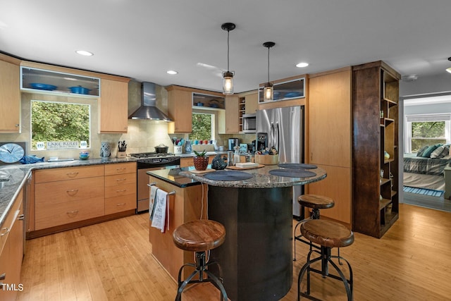 kitchen featuring tasteful backsplash, light hardwood / wood-style flooring, a kitchen bar, appliances with stainless steel finishes, and wall chimney range hood