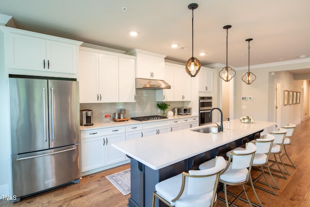 kitchen with appliances with stainless steel finishes, a kitchen island with sink, light hardwood / wood-style floors, hanging light fixtures, and sink