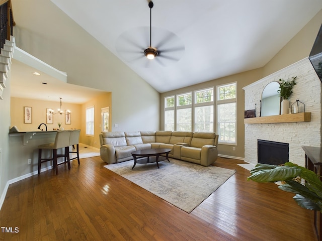 living room with dark wood-type flooring, ceiling fan with notable chandelier, a stone fireplace, and high vaulted ceiling