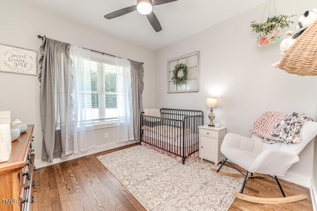 bedroom with baseboards, wood finished floors, ceiling fan, and a nursery area
