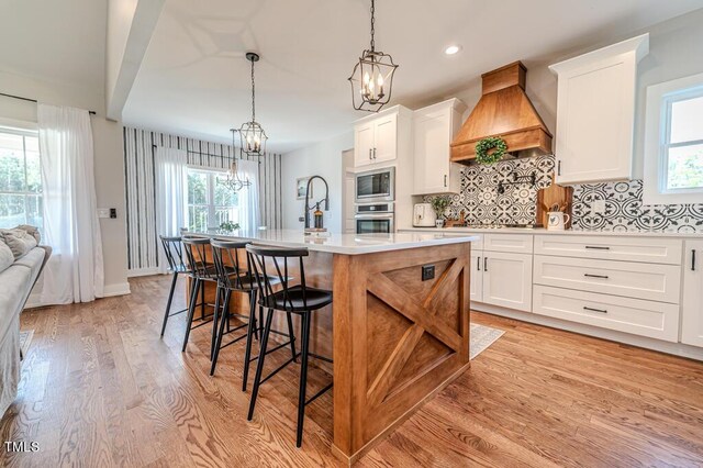 kitchen with premium range hood, light wood-style flooring, stainless steel appliances, a kitchen island with sink, and an inviting chandelier