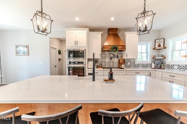 kitchen featuring light countertops, premium range hood, stainless steel appliances, backsplash, and a notable chandelier