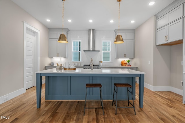 kitchen with wall chimney exhaust hood, hanging light fixtures, light wood-type flooring, and a center island with sink