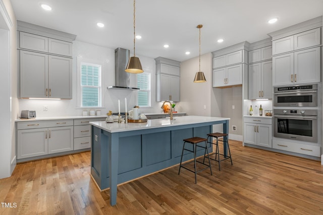 kitchen featuring a kitchen island with sink, wall chimney exhaust hood, decorative light fixtures, light wood-type flooring, and gray cabinets