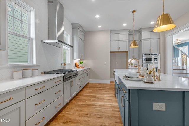 kitchen with a center island with sink, hanging light fixtures, appliances with stainless steel finishes, light wood-type flooring, and wall chimney exhaust hood