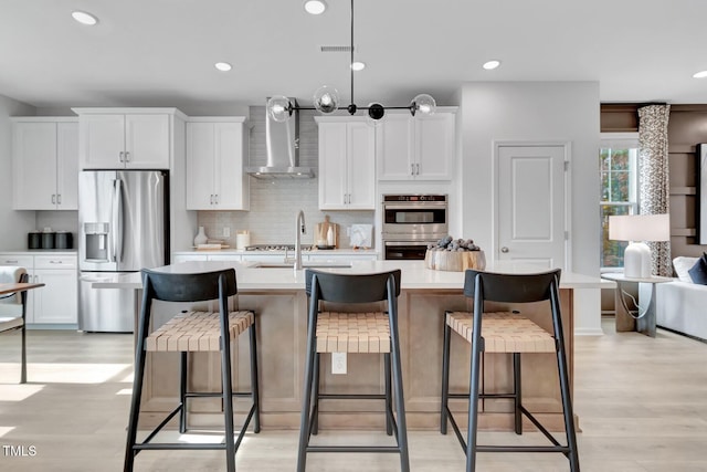 kitchen featuring stainless steel appliances, a center island with sink, wall chimney range hood, white cabinets, and a kitchen breakfast bar