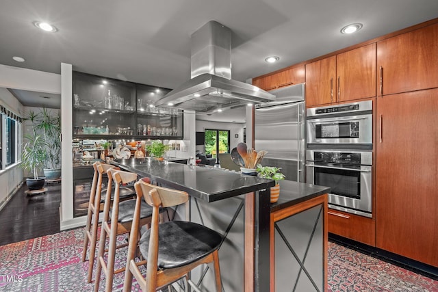 kitchen with island exhaust hood, recessed lighting, dark countertops, a kitchen bar, and stainless steel appliances