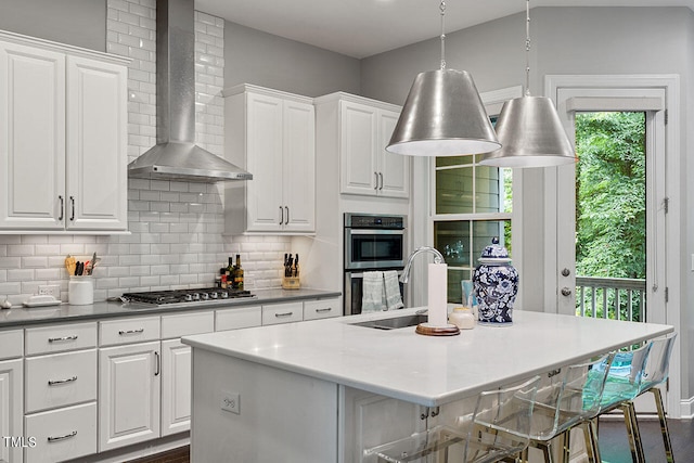 kitchen featuring a kitchen island with sink, white cabinetry, wall chimney exhaust hood, stainless steel appliances, and backsplash