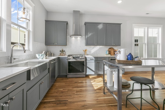 kitchen featuring gray cabinetry, hanging light fixtures, appliances with stainless steel finishes, wall chimney range hood, and dark hardwood / wood-style floors