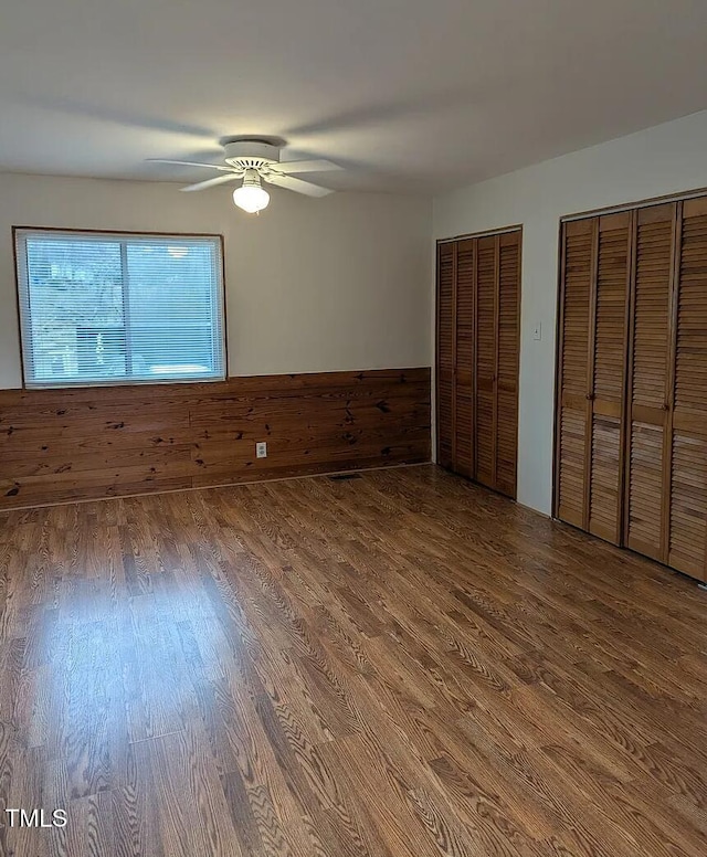 unfurnished bedroom featuring ceiling fan, a wainscoted wall, wood walls, wood finished floors, and multiple closets