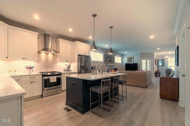 kitchen featuring appliances with stainless steel finishes, wall chimney exhaust hood, a center island with sink, decorative light fixtures, and white cabinetry