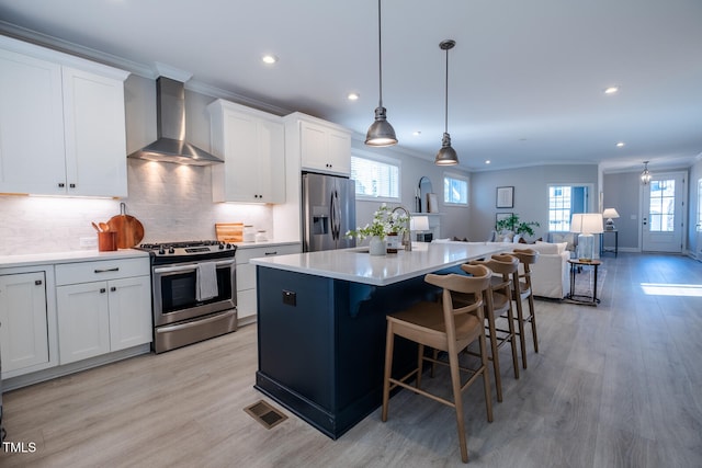 kitchen with a center island with sink, stainless steel appliances, light countertops, wall chimney range hood, and white cabinetry