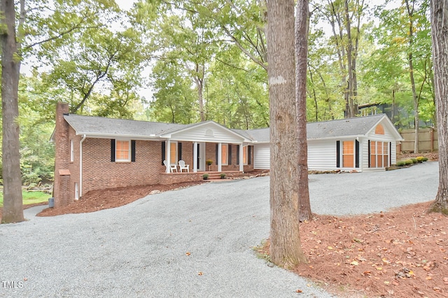 ranch-style house featuring covered porch, brick siding, a chimney, and gravel driveway